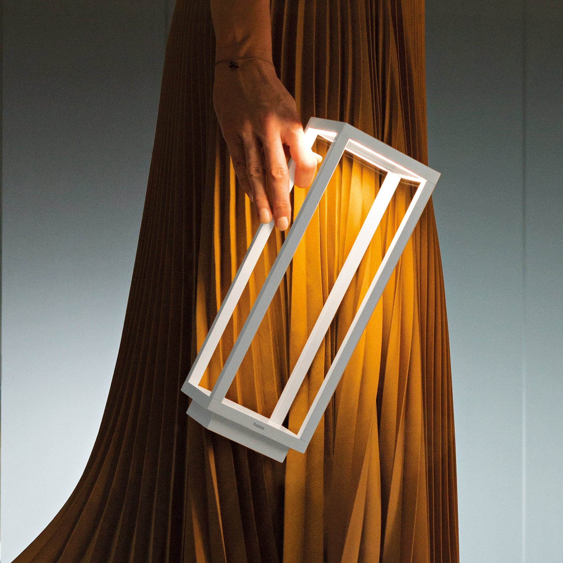 A person holds the Zafferano Home Pro Lamp in white, a rectangular minimalist rechargeable lamp, against pleated brown fabric.