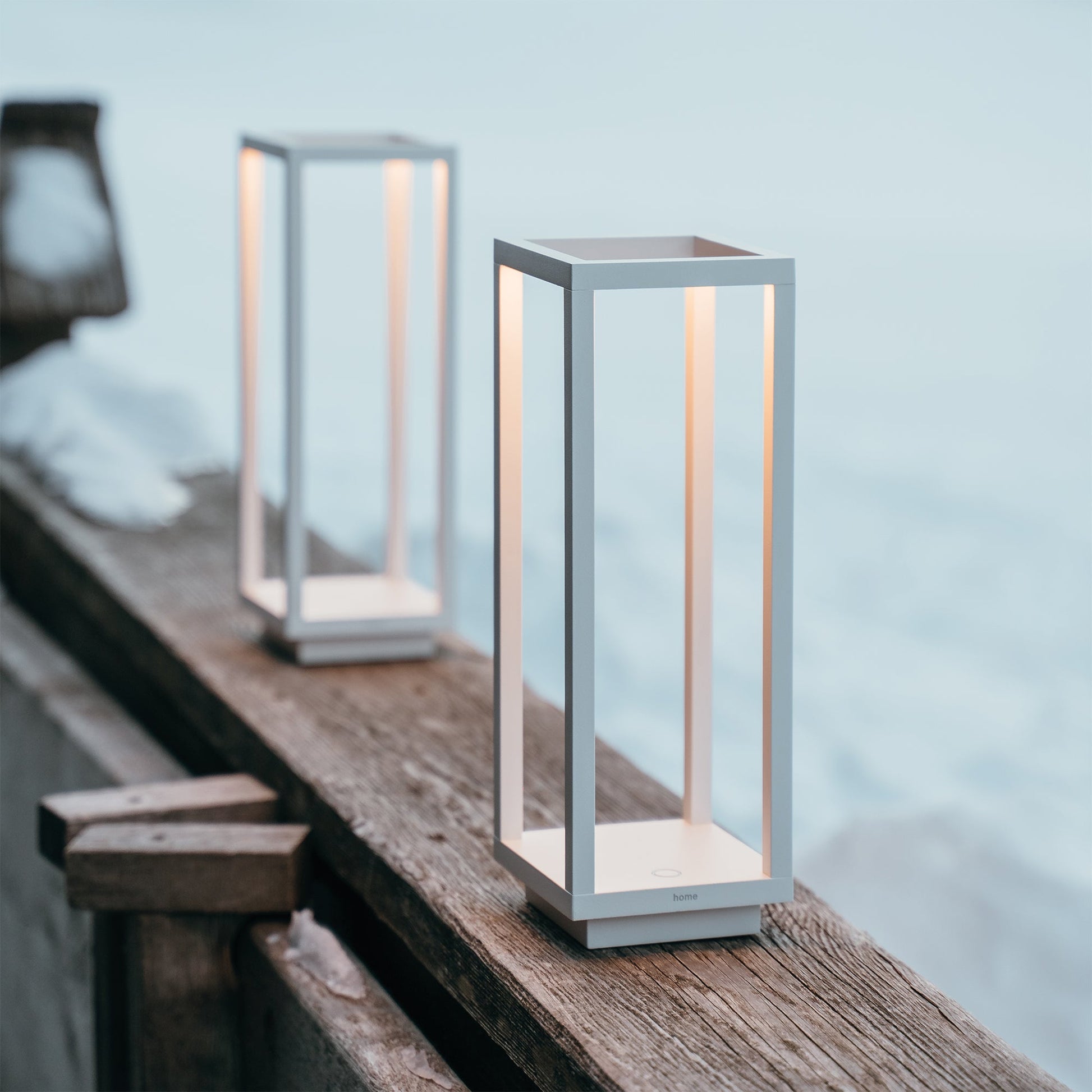 Two modern, rectangular Zafferano Home Pro Lamps in white emit a warm light on a wooden outdoor railing in the snow, combining minimalistic design with versatile indoor-outdoor cordless lighting.