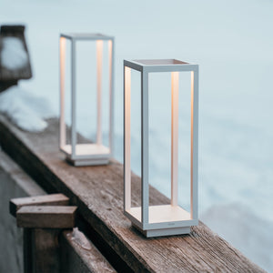 Two modern, rectangular Zafferano Home Pro Lamps in white emit a warm light on a wooden outdoor railing in the snow, combining minimalistic design with versatile indoor-outdoor cordless lighting.