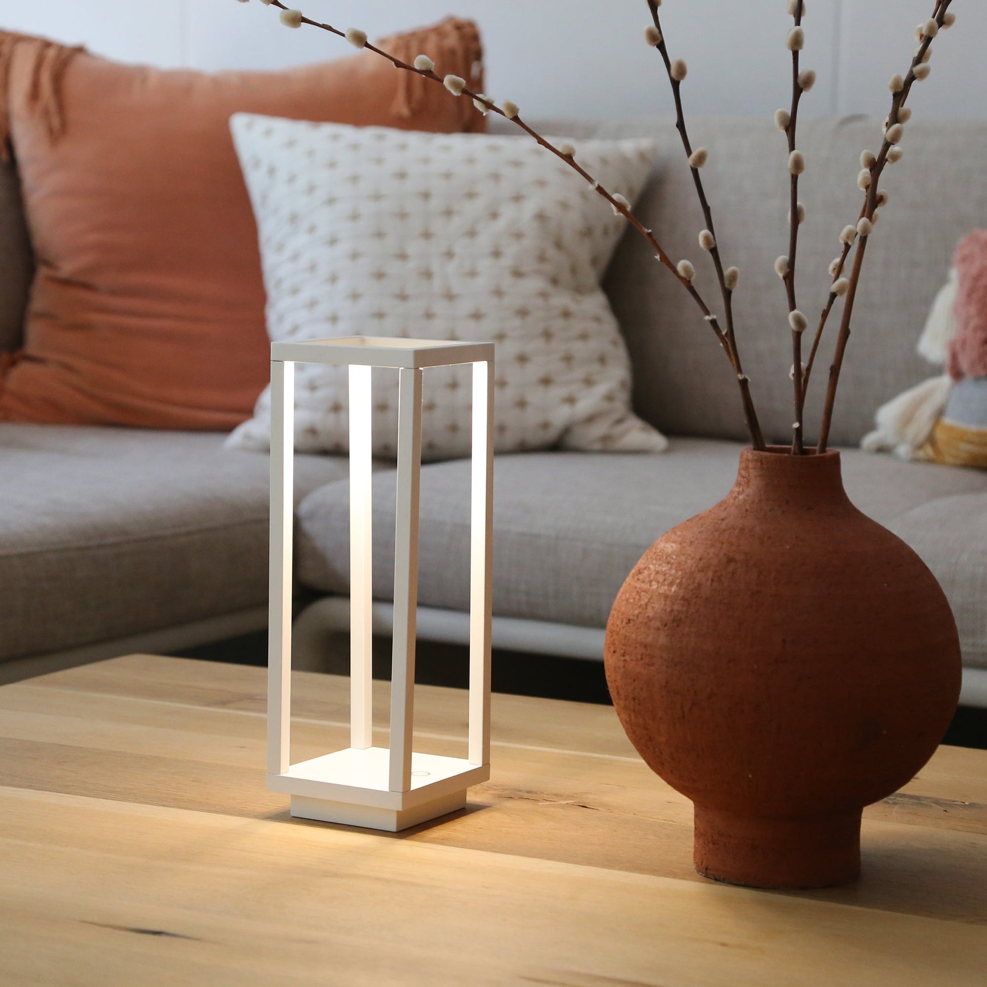 The Zafferano Home Pro Lamp, White, sits on a wooden coffee table beside a terracotta vase with branches, in front of a beige couch—perfect for stylish indoor or outdoor lighting.