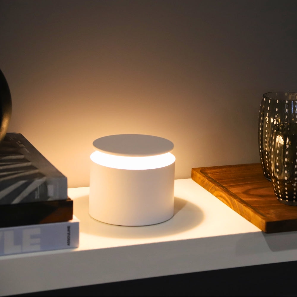 A compact, modern Zafferano PushUp Pro Lamp casts light on a shelf beside stacked books, a wooden tray, and a dark glass tumbler.