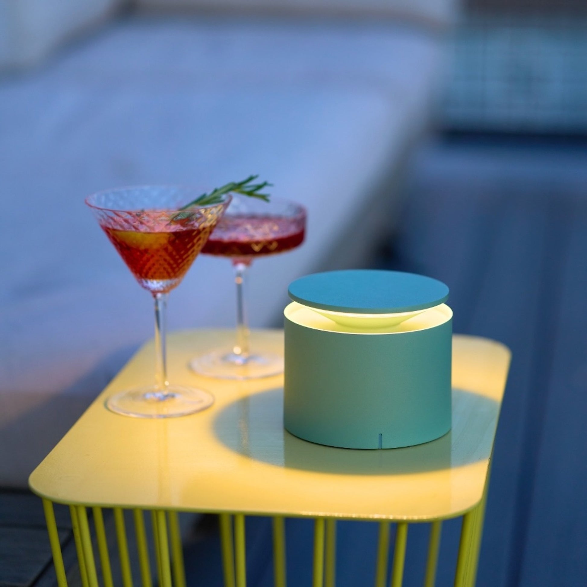 Two cocktails in stemmed glasses and a Zafferano PushUp Pro cordless table lamp rest on a small yellow table beside a sofa.
