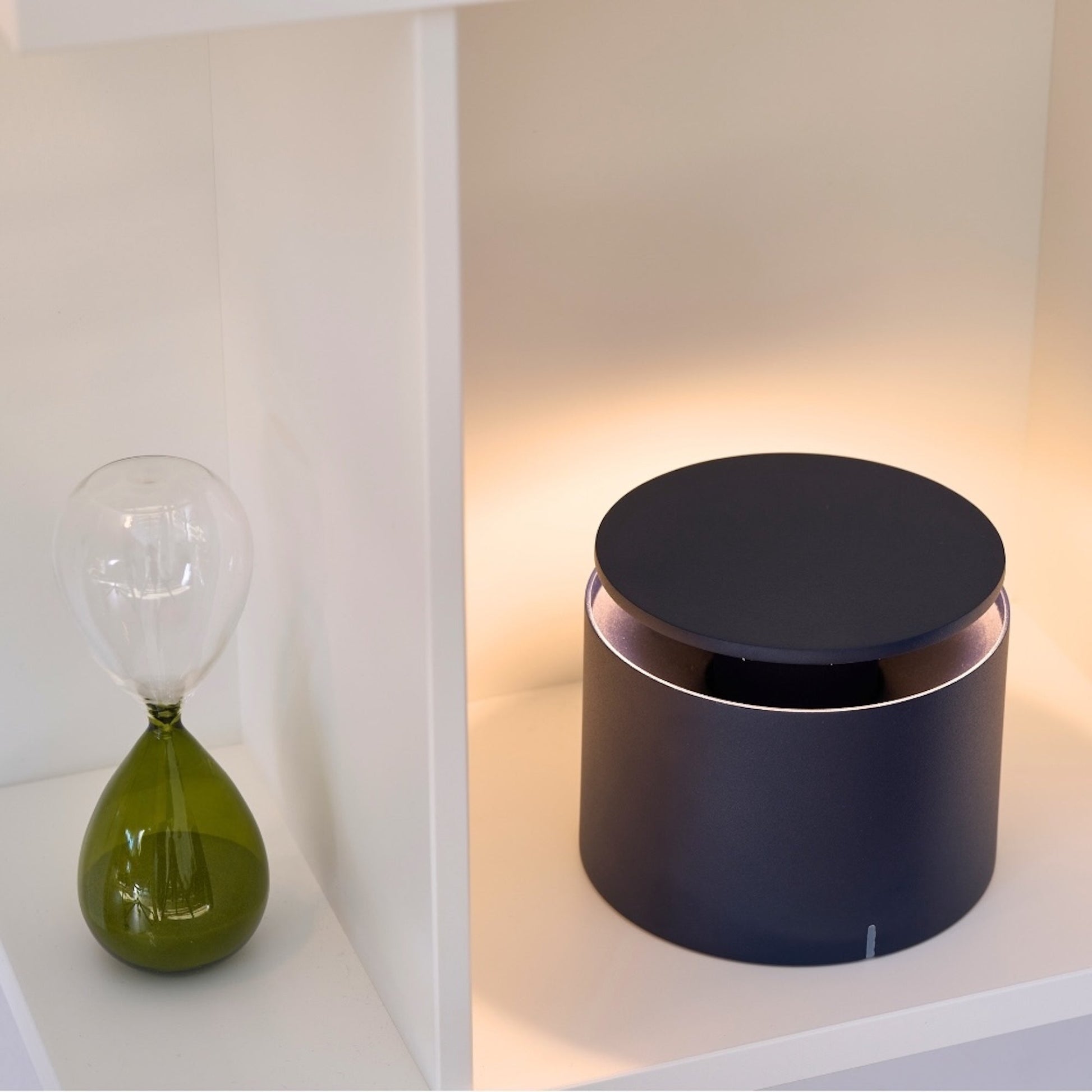A green hourglass and a Zafferano PushUp Pro Lamp, a round black rechargeable lamp from the collection, sit on separate white shelves while the lamp emits a soft light.