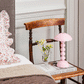 A wooden chair beside a bed holds an Addison Ross Bobbin LED Cordless Lamp, a small vase with white flowers, and a book titled "CURATE.