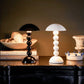 Two Addison Ross Bobbin LED Cordless Lamps—one black, one white—sit on a dark surface beside books, a bowl of shells, and a ceramic piece, highlighting the sleek elegance of these modern table lamps.