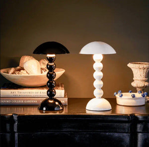 Two Addison Ross Bobbin LED Cordless Lamps—one black, one white—sit on a dark surface beside books, a bowl of shells, and a ceramic piece, highlighting the sleek elegance of these modern table lamps.