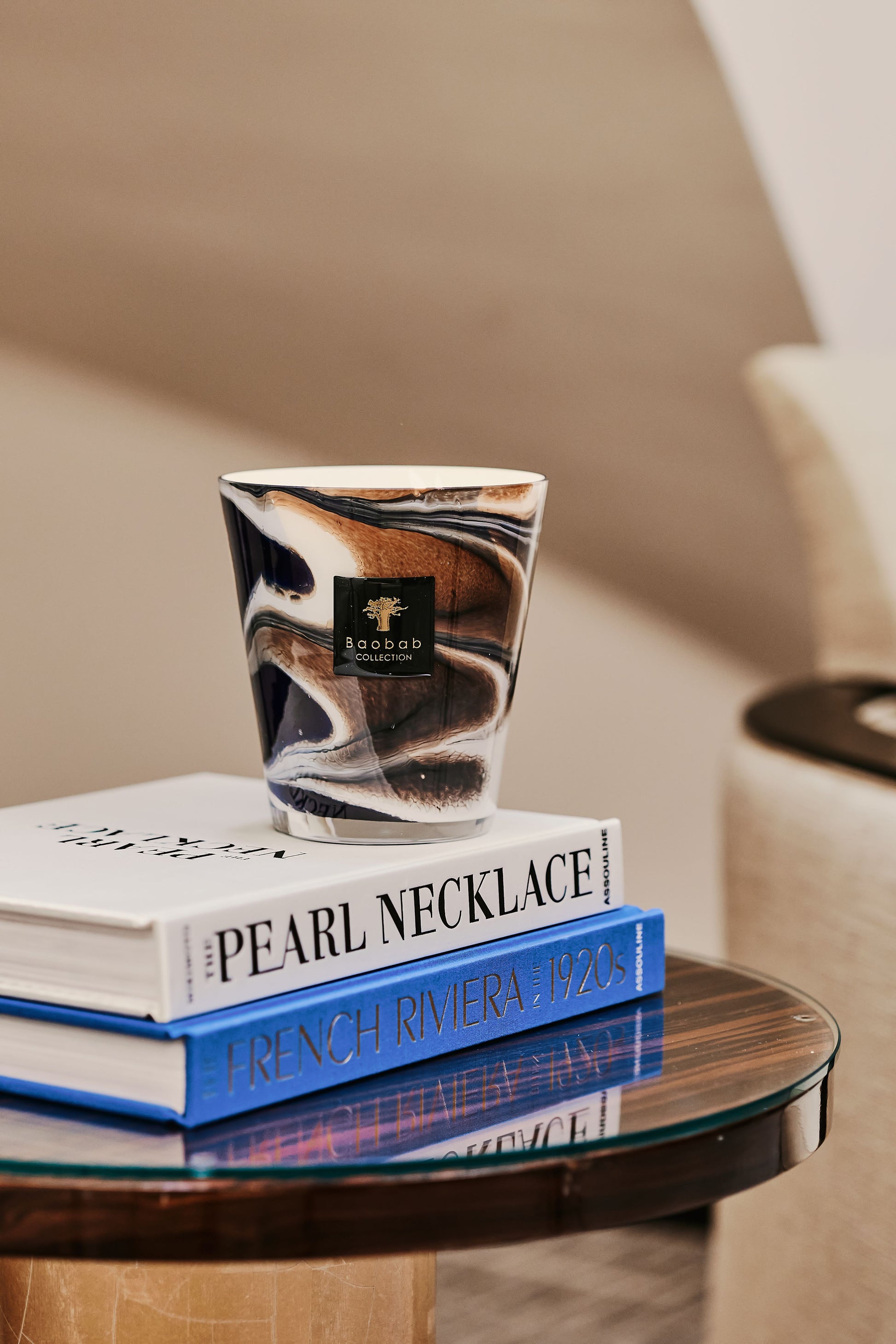 A marbled cup labeled "Baobab Collection Delta Nil Candle" rests on a stack of three books titled "Pearl Necklace," "French Riviera," and "1920s," its scent infused with Papyrus Blossom, all elegantly displayed on a small round glass table.