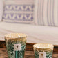 Two Baobab Collection Sacred Trees Kamalo Candles in decorative glass holders infused with cedarwood sit on a wooden surface, with a blurred background of a white couch and patterned cushions.