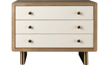 The Waterfall Nightstand is designed with a wooden dresser frame, featuring three white drawers adorned with brass knobs and supported by four short brass legs.