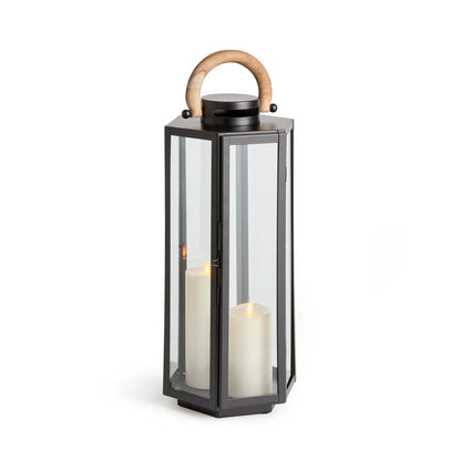 The Napa Dockside Lantern features a hexagonal black metal frame with clear glass panels, a natural teak wooden handle, and comes shown with two lit pillar candles inside.
