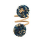 The Kim Seybert Casbah Napkin Ring displays organic elegance with a tan spiral coil and blue and beige yarn pom-poms on each end, shown isolated on a white background.