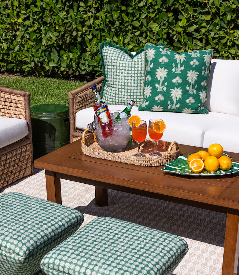 vignette of outdoor seating area with teak coffee table, woven sofa with white cushions and green patterened pillows