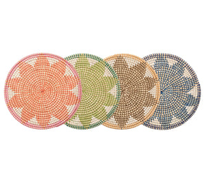 Four round placemats from the Kim Seybert Majorelle Placemat Collection are arranged side by side, displaying geometric patterns in pink, green, tan, and blue hues inspired by seagrass and subtle floral motifs.