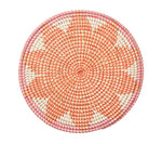 The Kim Seybert Majorelle Placemat Collection features round woven basket trays made from natural seagrass with a spiral orange, white, and pink pattern, white triangular accents, and a flat design, shown on a white background.