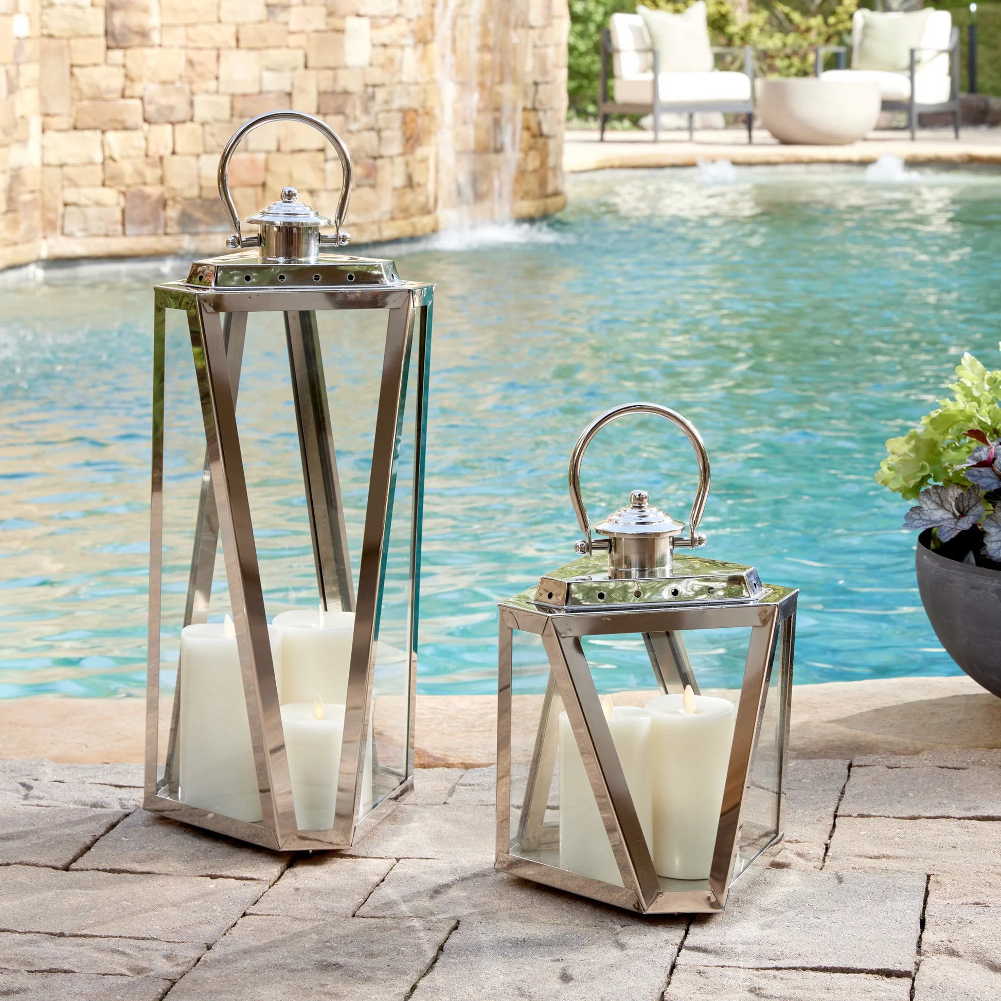 Two Napa Adler Steel Outdoor Lanterns with chrome finish and white LightLi pillars are placed on a stone patio beside a swimming pool.