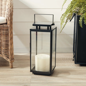 A Napa Alden Outdoor Lantern Small with black metal and glass frames a white pillar candle, bringing elegant outdoor lighting to a light wood floor next to a wicker chair and potted fern.
