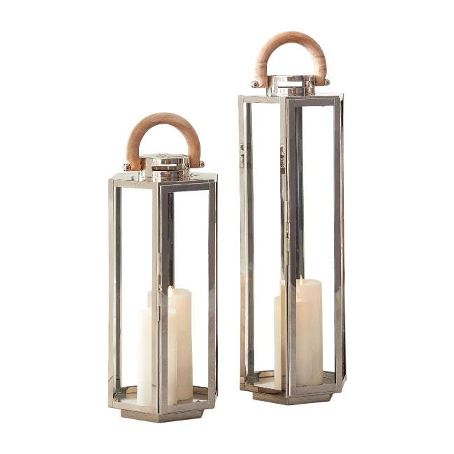 Two Dockside Lanterns, crafted from stainless steel and glass with natural teak handles, each hold a white pillar candle. The lanterns are shown against a plain background, with one taller than the other.