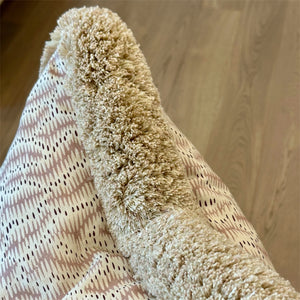 A close-up of a beige, fluffy blanket with a patterned edge on a light wooden floor—perfectly paired with Daphne's Feathers Rose Pillow, handcrafted from premium materials for added comfort and style.