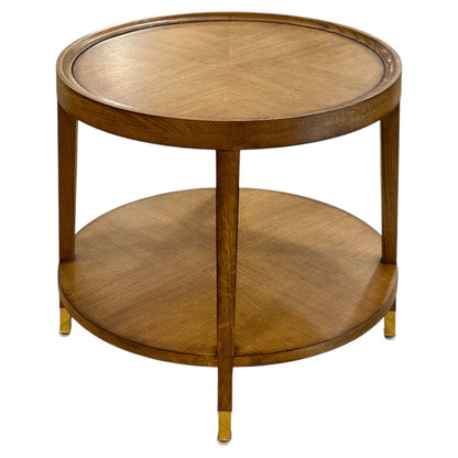 The Savoy Side Table, Wheat features a round wooden frame in a wheat finish, a lower shelf, and brass-capped feet for a simple, elegant look.