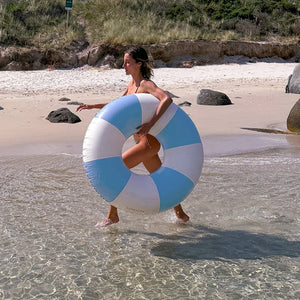 A woman carrying the SUNNYLiFE Poolside Tube Float Blue walks through shallow water on a sandy beach with grassy dunes, showcasing Australia’s summer style.
