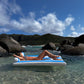 A person lounges on the SUNNYLiFE Luxe Lilo Blue Stripe inflatable in clear shallow water, embodying Australia’s summer style against rocky hills and a partly cloudy sky.