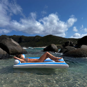 A person lounges on the SUNNYLiFE Luxe Lilo Blue Stripe inflatable in clear shallow water, embodying Australia’s summer style against rocky hills and a partly cloudy sky.