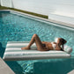 Enjoying the summer, a person in a white bikini relaxes on the SUNNYLiFE Luxe Lilo Green Stripe pool float, sunbathing in a clear swimming pool beside a white wall.