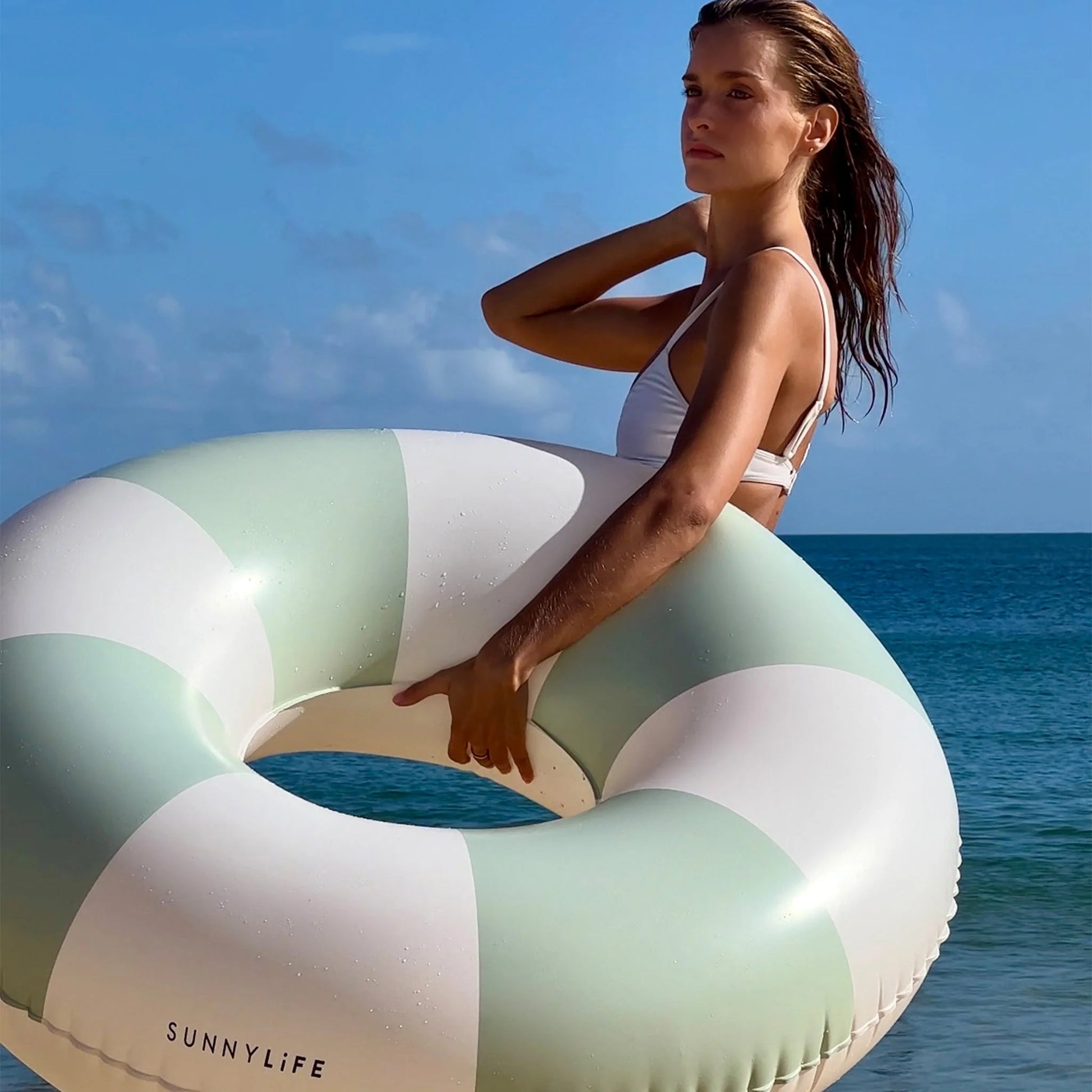 Woman in a white bikini stands on the beach with a SUNNYLiFE Poolside Tube Float Soft Olive, capturing Australia’s summer style against the ocean and blue sky—an iconic moment for this summer lifestyle brand.