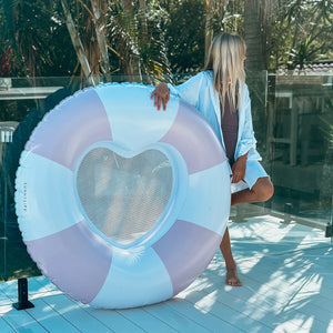 A person with long blond hair stands on a white deck, resting an arm on the SUNNYLiFE Luxe Poolside Tube Float Pink Stripe—its pastel stripes and heart-shaped center showcasing Australia’s summer style. Trees and glass fencing appear in the background.