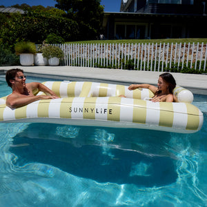 Two people relax on the SUNNYLiFE Luxe Twin Hammock Float Casa Sand Stripe, floating in a pool with a house and lush greenery in the background, capturing Australia’s summer style.