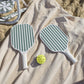 The SUNNYLiFE Pickle Ball Set in Olive, featuring green-and-white striped paddles with comfy foam handles and a yellow ball, sits on a beige towel in the sand by a folding chair—ideal for beachside fun.