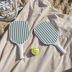 The SUNNYLiFE Pickle Ball Set in Olive, featuring green-and-white striped paddles with comfy foam handles and a yellow ball, sits on a beige towel in the sand by a folding chair—ideal for beachside fun.
