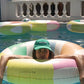 A person in a green cap leans on the SUNNYLiFE Poolside Tube Float Pastel Gelato in a swimming pool, capturing Australia’s summer style, with other colorful inflatables floating nearby.