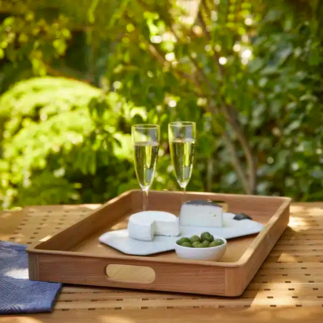 On a table, the SU Sundeck Teak Tray (22" x 17") holds two sparkling wine glasses, plates of two cheeses, and a small bowl of green olives, capturing yacht-inspired elegance with lush greenery in the background.