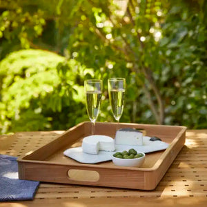 On a table, the SU Sundeck Teak Tray (22" x 17") holds two sparkling wine glasses, plates of two cheeses, and a small bowl of green olives, capturing yacht-inspired elegance with lush greenery in the background.