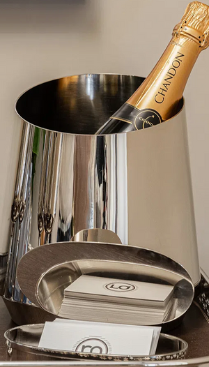 A bottle of Chandon champagne partially submerged in an ice bucket, elegantly decorating a St. James Silver Cachepot Geo Cooler next to a pile of business cards on a metal tray.