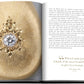 An open book displays an intricate gold brooch adorned with exquisite gems and a large central gemstone on the left page, while the right page features text including a quote and description about jewelry design in *Jewelry Guide: The Ultimate Compendium*.