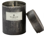 A cylindrical metal candle container labeled "Thucassi Ferrum Candle Collection, Leather Satchel." The lid is slightly removed and placed next to the container, hinting at notes of cured leather and red mandarin peel.