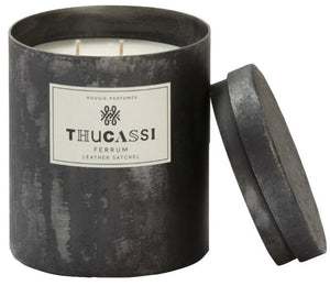 A black cylindrical candle with a lid, labeled "Thucassi Ferrum Candle Collection, Leather Satchel," featuring two wicks and the warm aroma of cured leather.