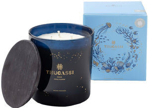 A blue glass candle container with a "Thucassi Ocean Candle Collection, Coral Garden" label, shown with its lid off beside a light blue box adorned with an ocean-themed design, emanates the refreshing scent of crisp Champagne grapes.