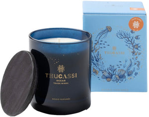 A Thucassi Ocean Candle Collection, Trade Winds scented candle in a blue glass jar with a black lid, infused with notes of bergamot and amber resin, next to its blue box featuring sea-themed illustrations.