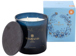 A blue Thucassi Ocean Candle Collection, Trade Winds scented candle with "Ocean Tides Wishes" written on the glass sits beside its decorative blue box featuring marine illustrations. The wax appears white and gives off hints of juniper berry, and the candle has a black lid.