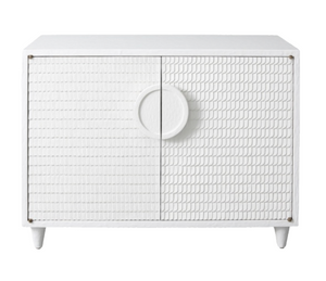 The Lance Cabinet is a white, hand-crafted piece featuring a textured, scale-like pattern on its double doors and a large circular handle in the center. This modern cabinet stands on four small legs, adding a minimalist accent to any room.