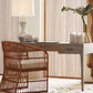 The Dublin Desk, a compact solid oak piece with a drawer, pairs with a wicker chair, lamp, vase of greenery, and decor on a cream rug against vertical wooden slats for a cozy yet functional workspace.