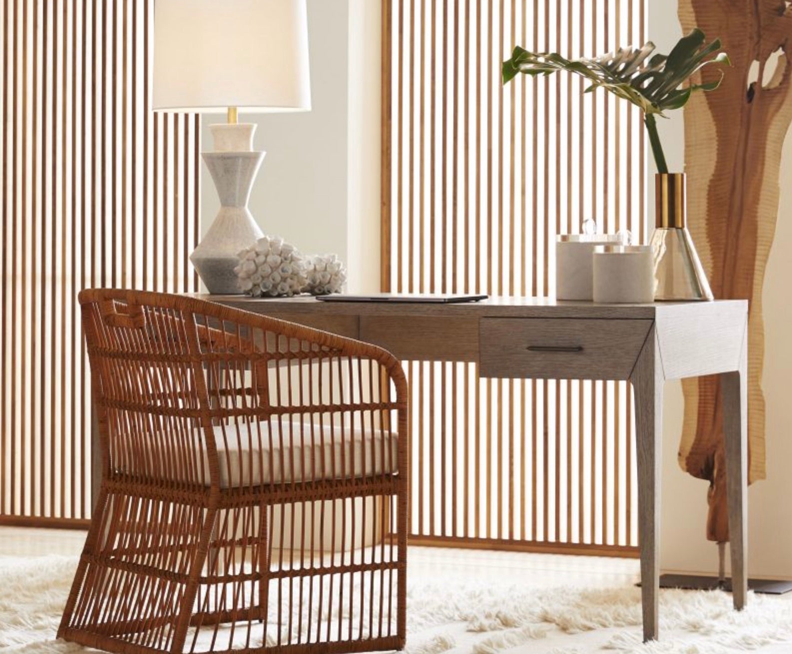The Dublin Desk, a compact solid oak piece with a drawer, pairs with a wicker chair, lamp, vase of greenery, and decor on a cream rug against vertical wooden slats for a cozy yet functional workspace.
