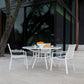 The Slant Outdoor Round Dining Table with four chairs sits on a wooden deck, ideal for garden gatherings. Bowls of oranges, a cup, and saucers are on the table, with a modern glass building and tree in the background.
