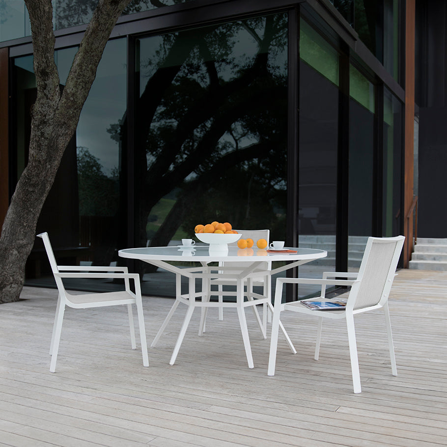 The Slant Outdoor Round Dining Table with four chairs sits on a wooden deck, ideal for garden gatherings. Bowls of oranges, a cup, and saucers are on the table, with a modern glass building and tree in the background.
