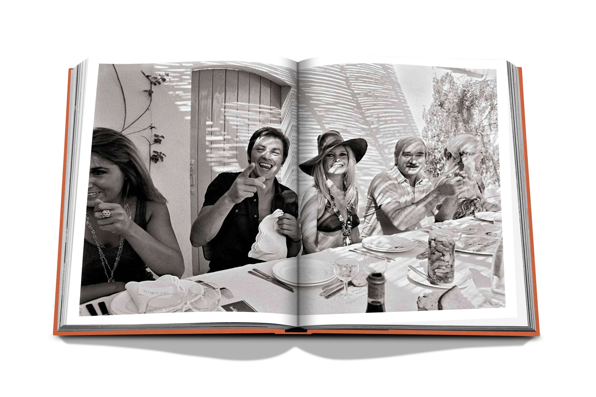 A black-and-white photograph in an open book shows a group of people at a sunlit outdoor table in St. Tropez, smiling and laughing. The table is set with plates, glasses, and a bottle, capturing the essence of St Tropez Soleil.