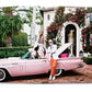 A woman sits on the back of a pink vintage car with the trunk open, surrounded by greenery and a building reminiscent of Star Style: Interiors of Martyn Lawrence Bullard. A man in an orange suit jacket and white pants stands beside the car.