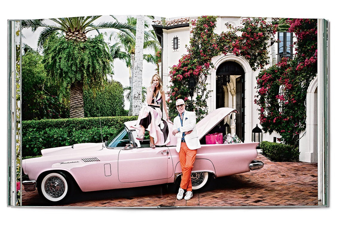 A woman sits on the back of a pink vintage car with the trunk open, surrounded by greenery and a building reminiscent of Star Style: Interiors of Martyn Lawrence Bullard. A man in an orange suit jacket and white pants stands beside the car.